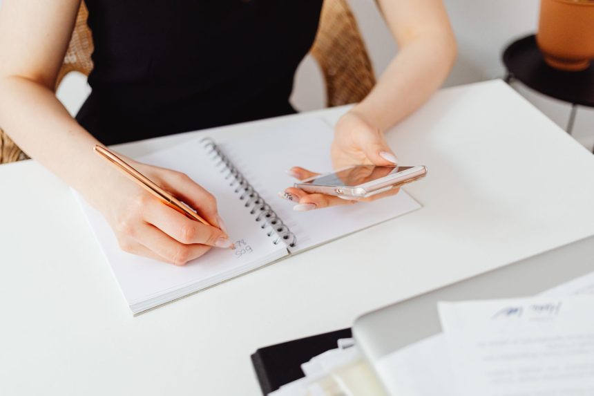 Mulher escreve em um caderno enquanto segura um smartphone, representando a organização de várias tarefas.