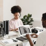 Dois profissionais criativos colaboram em um escritório moderno, usando laptops ao lado de equipamentos de câmera.