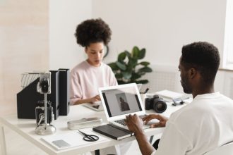 Dois profissionais criativos colaboram em um escritório moderno, usando laptops ao lado de equipamentos de câmera.