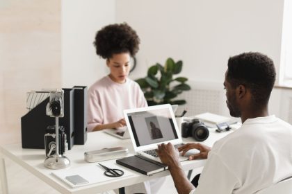 Dois profissionais criativos colaboram em um escritório moderno, usando laptops ao lado de equipamentos de câmera.