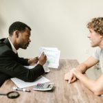 Profissional financeiro conversa com cliente enquanto analisa documentos em uma mesa de escritório.